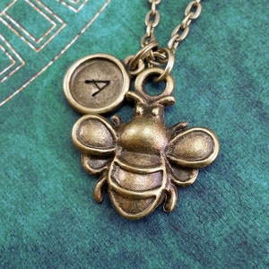 May include: A gold-toned necklace with a bee charm and a round charm with the letter 'A' engraved on it.