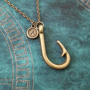 May include: A gold-toned necklace with a small charm that says "A" and a large hook pendant. The necklace is on a teal background.