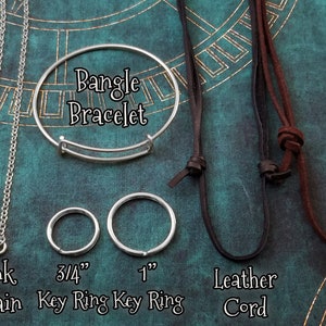 May include: A close-up of different types of jewellery chains and cords. The chains include a ball chain, a link chain, and a bangle bracelet. The cords include a leather cord and a suede cord. The chains and cords are labelled with their names and sizes. Bangle Bracelet, Ball Chain, Link Chain, 3/4" Key Ring, 1" Key Ring, Leather Cord, Suede Cord.