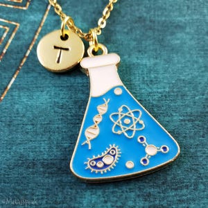 May include: Gold-tone necklace with a science-themed pendant. The pendant is a blue flask with white accents and scientific symbols. A gold disc with the letter "T" hangs next to the flask. The necklace is on a teal background.