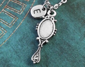 May include: A silver necklace with a hand mirror pendant and a charm with the letter "E". The mirror pendant is detailed. The necklace is shown on a teal textured background.