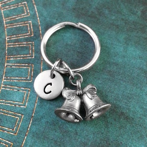 May include: Silver keychain with two small bells and a charm with the letter C. The keychain is on a teal background.