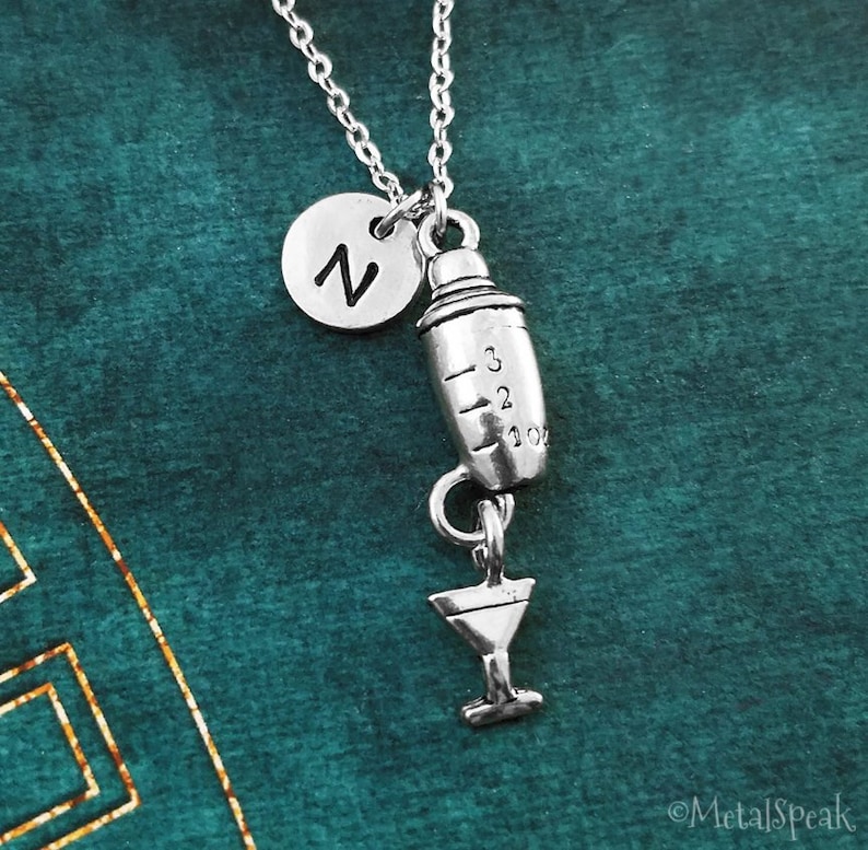 May include: Silver necklace with a silver cocktail shaker charm and a silver martini glass charm. The shaker charm has measurements on it. The necklace also has a silver initial charm with the letter "Z" on it.