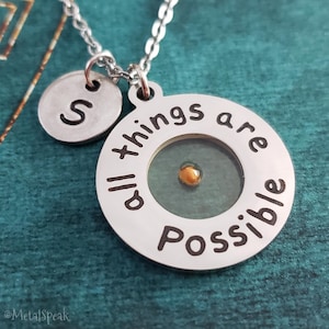 Könnte beinhalten: Silberfarbene Halskette mit einem runden Anhänger mit der Aufschrift "All things are possible". Der Anhänger hat einen kleinen Senfkorn in der Mitte. Ein zusätzlicher runder Anhänger mit dem Buchstaben "S" hängt daneben.