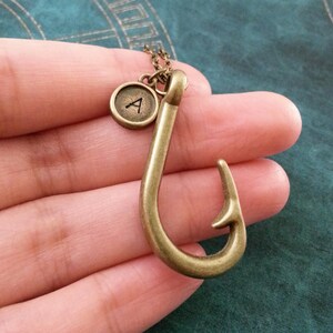 May include: A gold-toned necklace with a fish hook pendant and a small round charm with the letter 'A' on it.