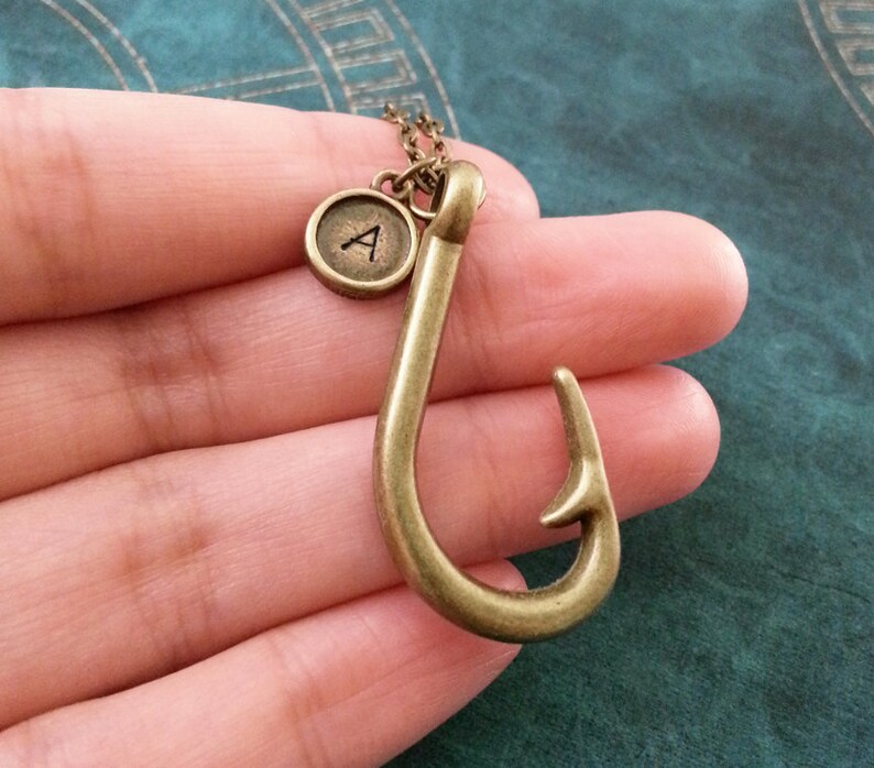 May include: A gold-toned necklace with a fish hook pendant and a small round charm with the letter 'A' on it.