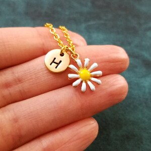 Pu&ograve; includere: Collana con catena dorata con un piccolo ciondolo a margherita bianca e un ciondolo a disco dorato con la lettera H.