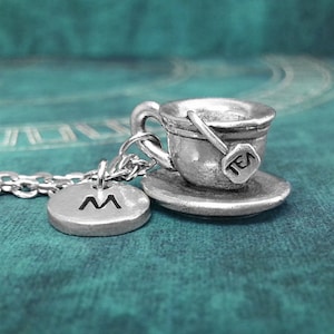 May include: A silver necklace with a charm of a teacup and saucer. The teacup has a tea bag charm that says "TEA". The necklace also has a silver charm with the letter "M" on it.
