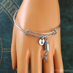 May include: Silver charm bracelet featuring a cocktail shaker and martini glass charm. The bracelet also has a personalised charm with the letter "R".