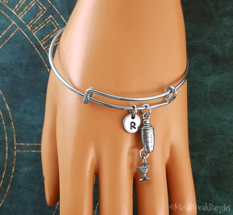 May include: Silver charm bracelet featuring a cocktail shaker and martini glass charm. The bracelet also has a personalised charm with the letter "R".