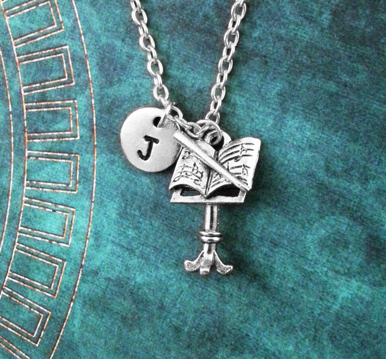 May include: Silver necklace with a music note charm, a music stand charm, and a letter J charm. The necklace is on a teal background.