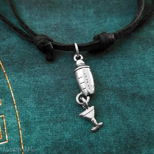 May include: A silver cocktail shaker charm with a martini glass charm hanging from it. The charms are attached to a black leather cord necklace.