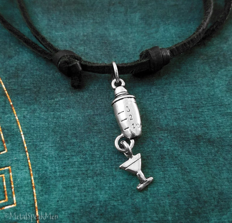May include: A silver cocktail shaker charm with a martini glass charm hanging from it. The charms are attached to a black leather cord necklace.