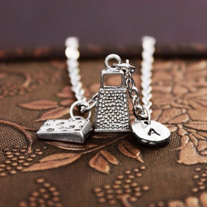 May include: A silver necklace with three charms: a cheese block, a grater, and a letter 'A'.
