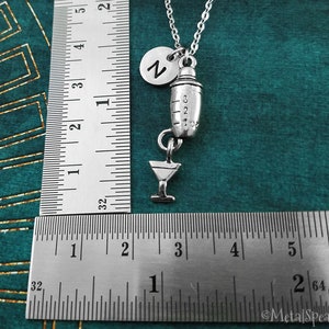 May include: Silver cocktail shaker charm necklace with a martini glass charm and a letter 'Z' charm. The necklace is on a silver chain and is shown against a green background.