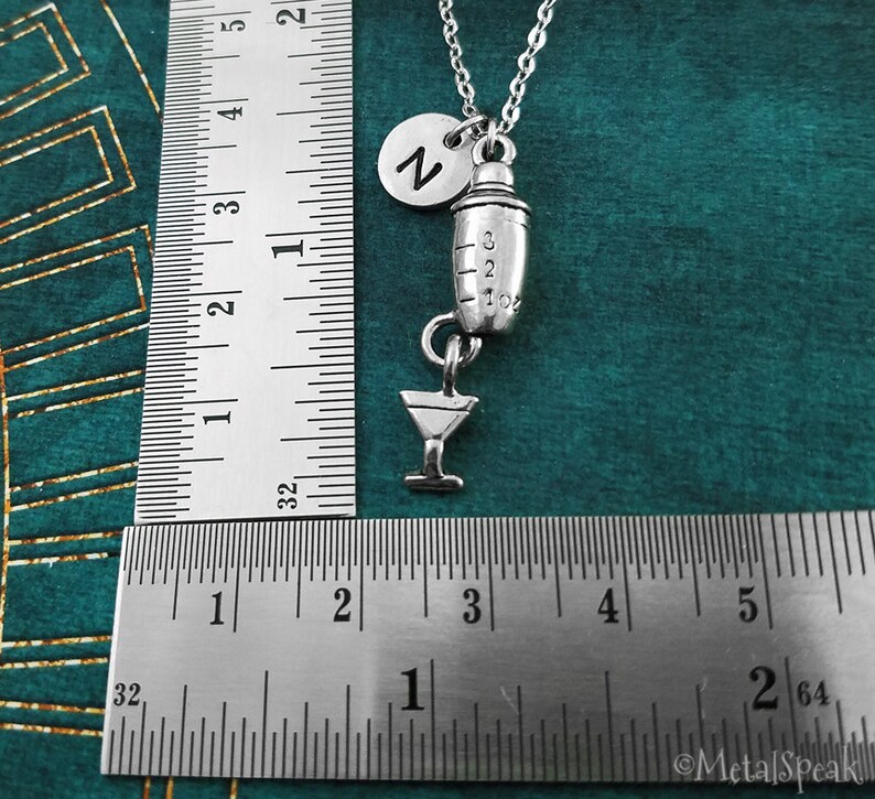 May include: Silver cocktail shaker charm necklace with a martini glass charm and a letter 'Z' charm. The necklace is on a silver chain and is shown against a green background.