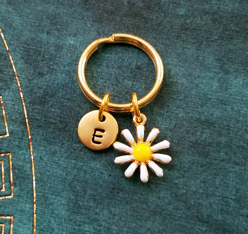 Pu&ograve; includere: Portachiavi dorato con un ciondolo a forma di margherita bianca e un ciondolo a disco dorato con la lettera "E" incisa.
