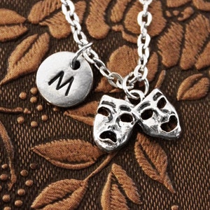 May include: Silver necklace with a silver charm that has the letter M on it. The charm is attached to two silver charms that are shaped like theater masks. One mask is smiling and the other is frowning.