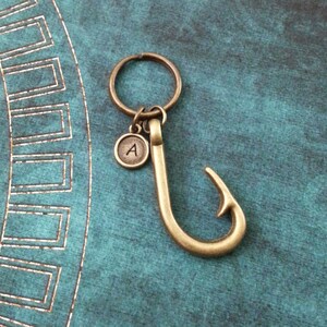 May include: A gold-toned keychain with a fish hook charm and a small round charm with the letter 'A' engraved on it.