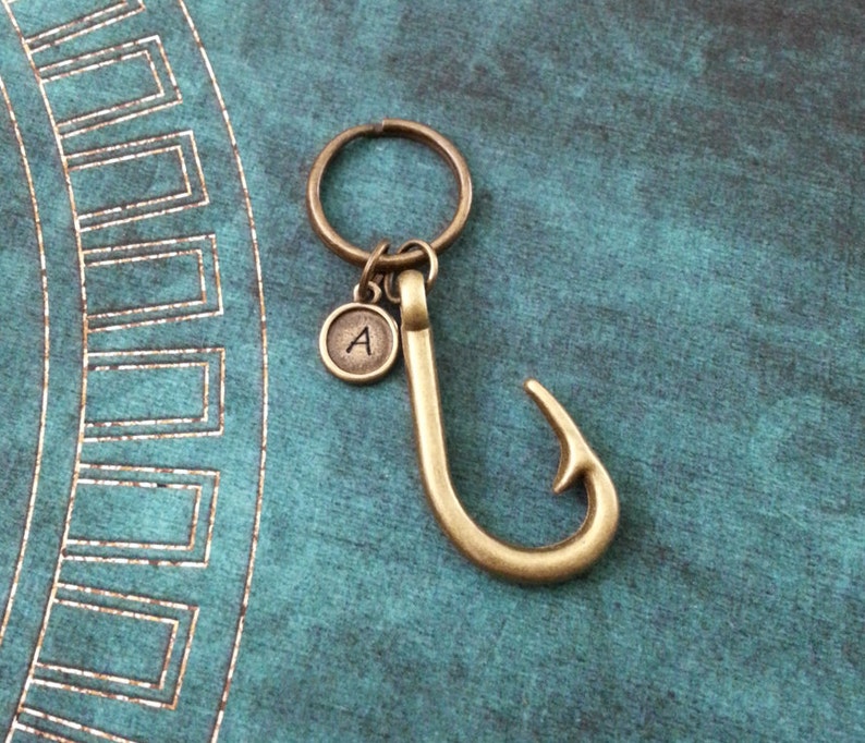 May include: A gold-toned keychain with a fish hook charm and a small round charm with the letter 'A' engraved on it.