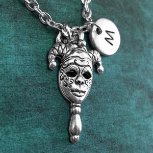May include: A silver necklace with a jester mask pendant and a silver charm with the letter 'M' on it. The necklace is on a teal background.
