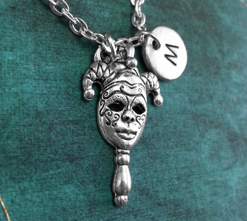 May include: A silver necklace with a jester mask pendant and a silver charm with the letter 'M' on it. The necklace is on a teal background.