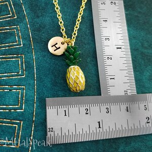 May include: Gold pineapple charm necklace with a gold initial "H" charm. The necklace is measured against a ruler.