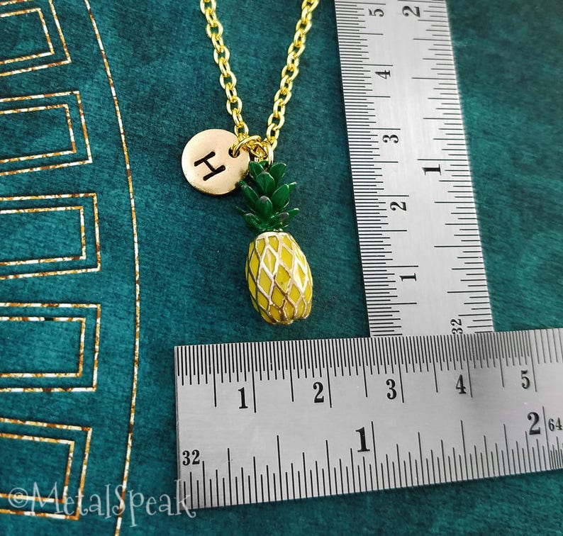 May include: Gold pineapple charm necklace with a gold initial "H" charm. The necklace is measured against a ruler.