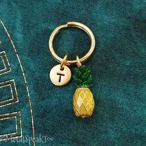 May include: Gold pineapple keyring with a letter 'T' charm. The pineapple is yellow and green with a gold ring.