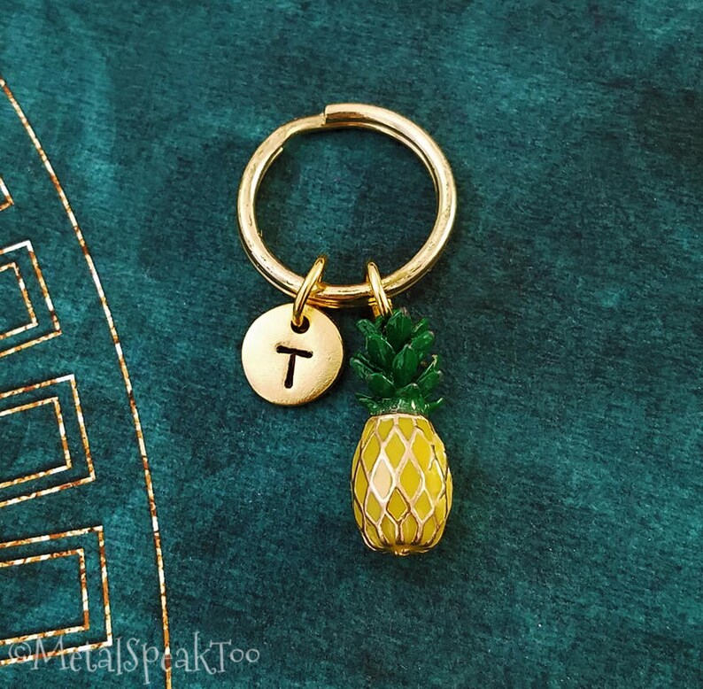 May include: Gold pineapple keyring with a letter 'T' charm. The pineapple is yellow and green with a gold ring.