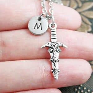 May include: Silver necklace with a sword charm and a round charm with the letter "M" on it.