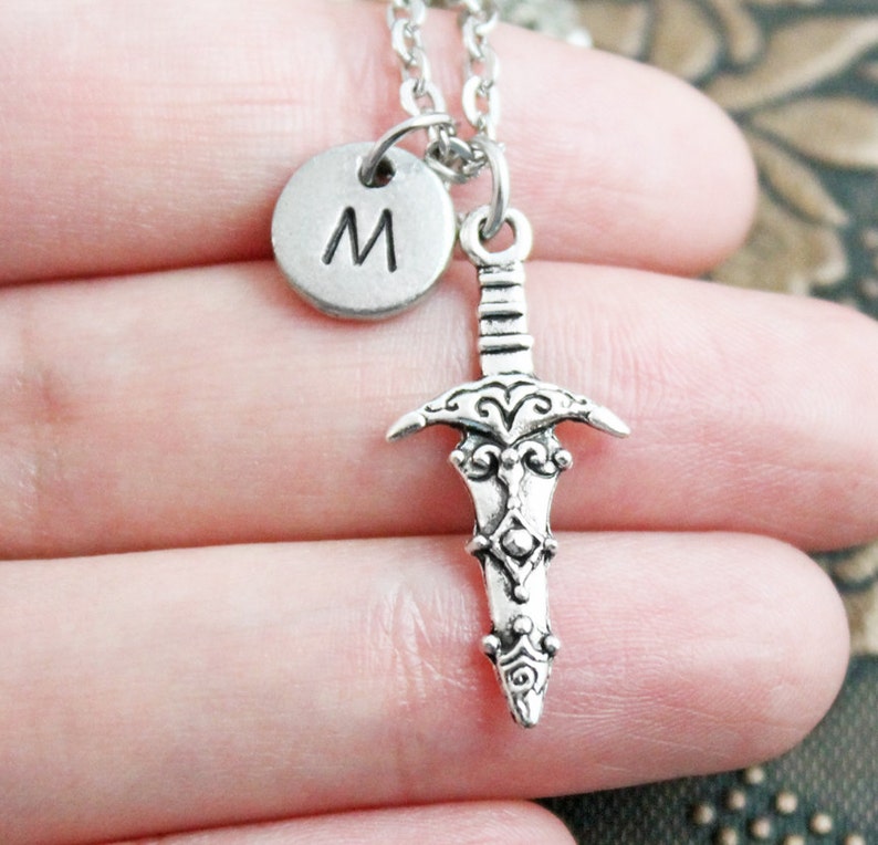 May include: Silver necklace with a sword charm and a round charm with the letter "M" on it.