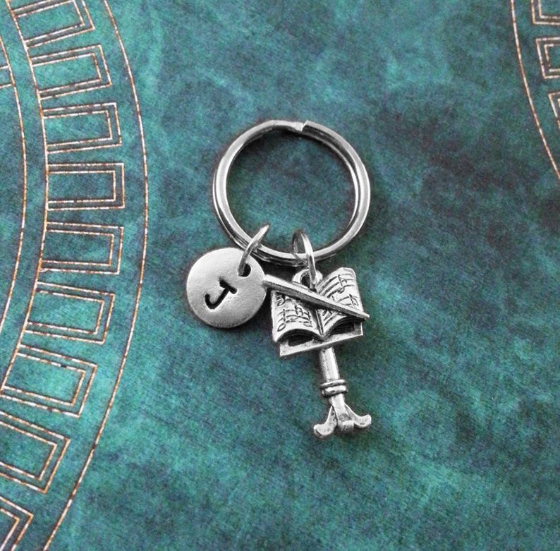 May include: Silver keychain with a music note book charm, a conductor's baton charm, and a letter J charm. The charms are attached to a silver key ring.