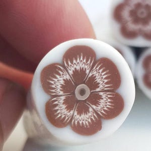 May include: Close-up of a white and brown floral bead. The bead features a brown flower design with white accents and a small central hole. The flower has five petals with a detailed, textured pattern.