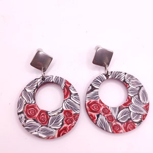 Può includere: Un paio di orecchini rotondi in argento e rosso con fiori e un ciondolo quadrato in argento. Gli orecchini hanno uno sfondo bianco con rose rosse e foglie grigie.