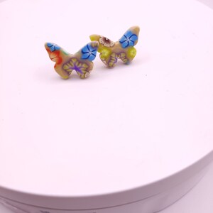 May include: A pair of butterfly-shaped earrings with a floral pattern in shades of yellow, blue, red, and green. The earrings are made of a light beige material.