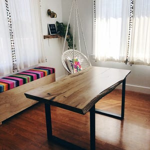 Op de afbeelding: Een lichte houten tafel met een levende rand en zwarte metalen poten. De tafel staat in een kamer met witte gordijnen en een hangende macramé stoel.
