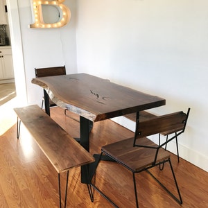 Op de afbeelding: Een houten eettafel met een live edge blad en zwarte metalen poten. De tafel is voorzien van twee stoelen en een bank, allemaal gemaakt van hout met zwarte metalen poten. De stoelen hebben een geometrisch ontwerp. De tafel staat in een kamer met een witte muur en een lamp in de vorm van de letter "B".