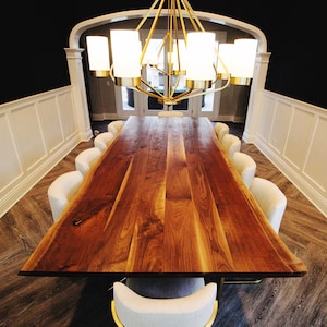 May include: A long, wooden dining table with a live edge design. The table is made of walnut wood and has a natural finish. The table is surrounded by white chairs and a gold chandelier hangs above.