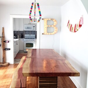 Op de afbeelding: Een grote eettafel van levend hout met een natuurlijke roodbruine afwerking. De tafel heeft een unieke, onregelmatige vorm en wordt ondersteund door een set gestapelte houten blokken. Een bijpassende bank staat links van de tafel. De tafel staat in een lichte, witte kamer met een kleurrijke, gestreepte hanglamp erboven.