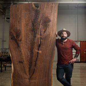 May include: A man wearing a brown hat and a red long-sleeved shirt stands next to a large slab of wood. The wood has a natural, dark brown color and a unique grain pattern. The wood is likely walnut and has been stabilized with butterfly keys.