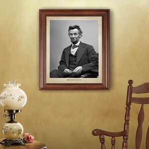 Puede incluir: Un retrato en blanco y negro enmarcado de Abraham Lincoln con traje y corbata. El marco es de madera marr&oacute;n con un borde dorado. El retrato est&aacute; colgado en una pared con una mecedora y una l&aacute;mpara en primer plano.