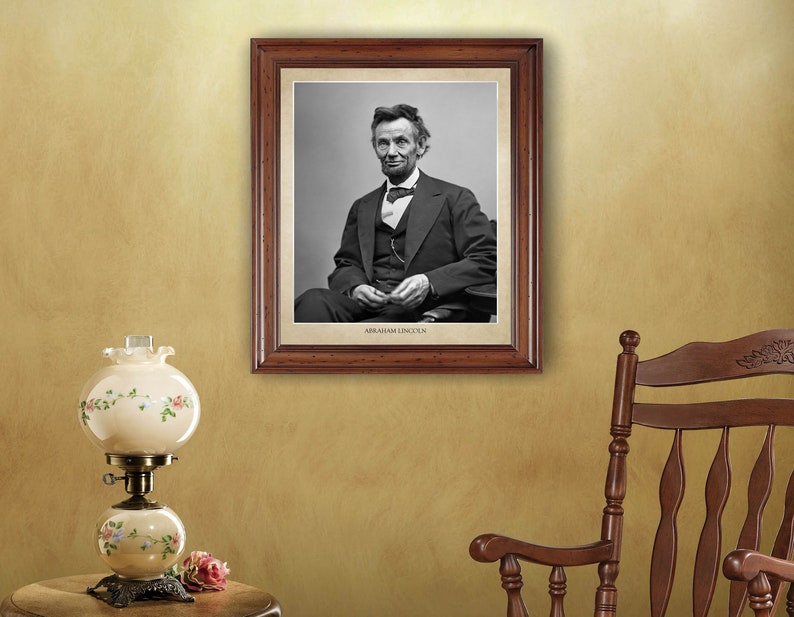 Puede incluir: Un retrato en blanco y negro enmarcado de Abraham Lincoln con traje y corbata. El marco es de madera marr&oacute;n con un borde dorado. El retrato est&aacute; colgado en una pared con una mecedora y una l&aacute;mpara en primer plano.