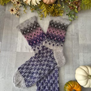 May include: A pair of hand-knitted wool socks featuring a grey, purple, and dark pink Fair Isle pattern on the cuffs and a textured purple and grey geometric pattern on the feet, finished with solid grey toes and heels.
