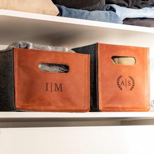 May include: Two rectangular storage bins made of dark gray felt with tan leather front panels. Each bin features a cutout handle and a personalized monogram. The left bin displays the letters I | M, while the right bin shows A | S inside a wreath.