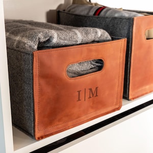 Peut inclure: Un bac de rangement rectangulaire en feutre gris foncé avec un panneau avant en cuir beige doté d'une poignée découpée et des lettres gravées I | M. Le bac contient une couverture à carreaux gris et blanc, offrant une solution de rangement.