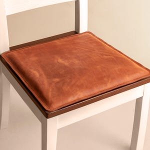 May include: A square, cognac-colored leather seat cushion with visible stitching along the edges. The cushion is placed flat on the dark brown wooden seat of a white chair, featuring a smooth, matte texture with natural variations in the leather.