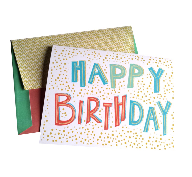 May include: A birthday card with the words "HAPPY BIRTHDAY" in blue, orange, and green. The card has a white background with gold polka dots. The envelope is green, red, and gold.