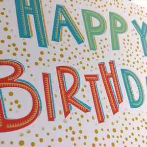 May include: A white birthday card featuring the words "HAPPY BIRTHDAY" in bold, colourful lettering. The letters are outlined in blue, red, and gold, set against a background of gold dots. Ideal for a birthday celebration.