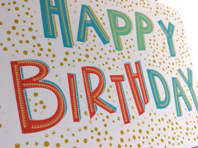 May include: A white birthday card featuring the words "HAPPY BIRTHDAY" in bold, colourful lettering. The letters are outlined in blue, red, and gold, set against a background of gold dots. Ideal for a birthday celebration.